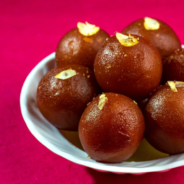 A white bowl filled with several glistening gulab jamun, garnished with slivered almonds, on a pink background.