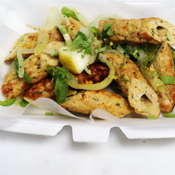 Indian seekh kebabs with sliced onions, cilantro, and a lemon wedge in a white container.