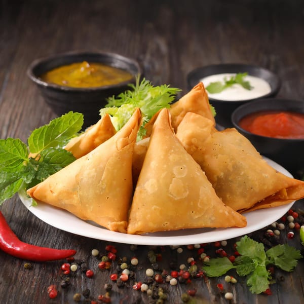 Golden-brown Indian samosas on a white plate with mint, served with three dipping sauces.
